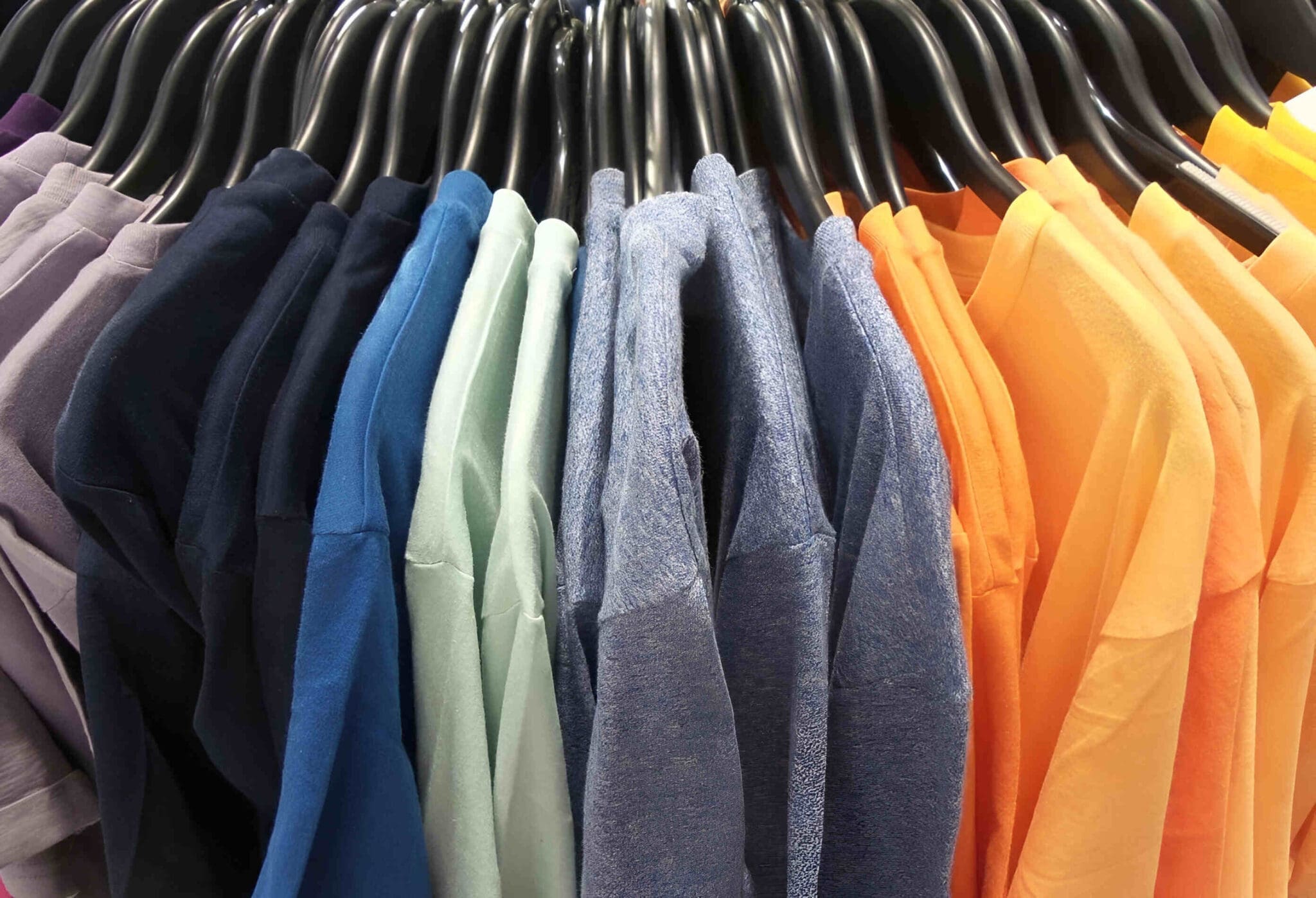 A row of assorted colored t-shirts, including shades of grey, black, blue, green, and orange, hanging on black hangers.