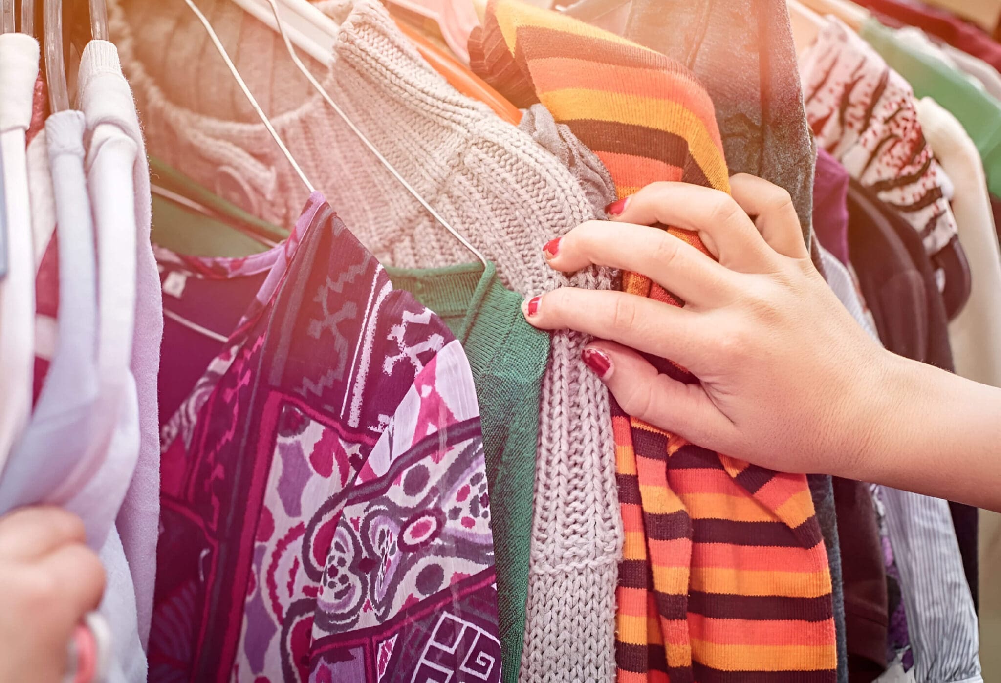 Close-up of a person's hand browsing through a rack of colorful clothing, including patterned shirts and sweaters on hangers.